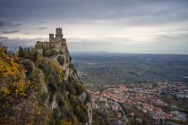Antiguo castillo en el pico cubierto de árboles verdes que se elevan alto en el cielo gris en San Marino, Italia - foto de stock