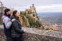 Hombre amoroso abrazando sonriente mujer asiática en sombrero de punto con pompón apoyado en valla de piedra en la colina con increíble paisaje del antiguo castillo en San Marino, Italia - foto de stock