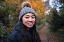 Giovane affascinante donna asiatica in giacca nera calda e cappello a maglia con pompon sorridente e guardando la fotocamera in strada verso il giardino con foglie autunnali a San Marino, Italia — Foto stock