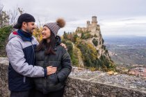 Homem amoroso abraçando sorridente mulher asiática em chapéu de malha com pompom inclinado sobre cerca de pedra na colina com paisagem incrível do castelo antigo em San Marino, Itália — Fotografia de Stock