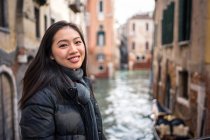 Zufriedene asiatische Frau im Urlaub in warmer Kleidung lächelt und blickt in die Kamera mit Wasserlinie zwischen alten Gebäuden auf verschwommenem Hintergrund — Stockfoto