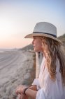 Side view of graceful woman with long curly hair in hat leaning on fence and looking away at seaside — Stock Photo