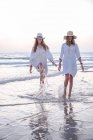 Smiling girlfriends in summer clothes barefoot in water on beach — Stock Photo