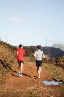 Vista trasera de hombres anónimos fuertes y activos en ropa deportiva corriendo juntos en el camino de tierra en las montañas en el soleado día de otoño - foto de stock