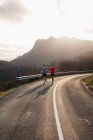 Corredores masculinos sanos activos que corren juntos en el camino curvo del asfalto con la luz del sol detrás de la montaña en el fondo - foto de stock