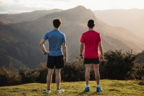 Vista posterior de los hombres deportivos en camisas azules y rojas de pie en la cima de una colina verde y disfrutando del paisaje mientras se relaja después de correr - foto de stock