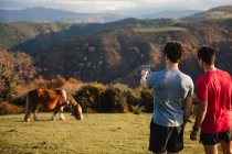 Vista posterior de los hombres deportivos de pie en la cima de la colina verde tomando fotos con el teléfono móvil de una vaca en el pasto - foto de stock
