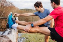 Vista lateral de joggers masculinos cansados em camisas azuis e vermelhas que se estendem na cerca de madeira depois de correr e treinar duro na colina verde — Fotografia de Stock