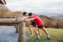 Side view of tired male joggers in blue and red shirts stretching on wooden fence after running and training hard on green hill — Stock Photo