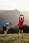 Rückenansicht müder männlicher Jogger in blauen und roten Hemden, die sich nach dem Laufen und hartem Training auf dem grünen Hügel strecken — Stockfoto