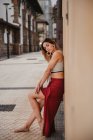 Side view of barefoot woman in trendy top and skirt leaning on building wall and looking at camera — Stock Photo