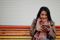 Азиатка с чемоданом сидит на радужной скамейке и просматривает смартфон на городской улице — стоковое фото