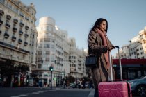Femme asiatique avec valise debout sur le trottoir près de la route et regardant loin tout en visitant la ville le jour ensoleillé — Photo de stock