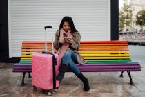 Asian woman with suitcase sitting on rainbow bench and browsing smartphone on city street — Stock Photo