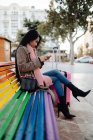 Side view Asian woman with suitcase sitting on rainbow bench and browsing smartphone on city street — Stock Photo