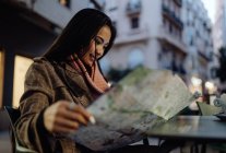 Joven mujer asiática sonriendo y leyendo mapa mientras se sienta a la mesa en la cafetería de la calle por la noche en la ciudad - foto de stock