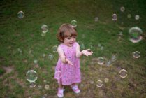 Зверху чарівна дитина в рожевому платті сміється і захоплює веселкові мильні бульбашки на зеленому лузі в парку — стокове фото