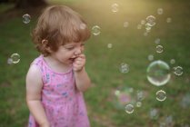 Сверху очаровательный ребенок в розовом платье смеется и захватывает радужные мыльные пузыри на зеленом лугу в парке — стоковое фото
