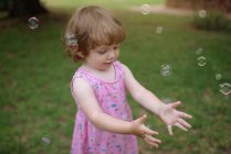 Зверху чарівна дитина в рожевому платті сміється і захоплює веселкові мильні бульбашки на зеленому лузі в парку — стокове фото
