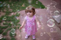 De cima criança adorável no vestido rosa rindo e capturando bolhas de sabão arco-íris no prado verde no parque — Fotografia de Stock