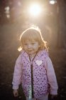 Чарівна весела дитина в теплому рожевому жилеті з серцем дивиться на камеру на сонячному світлі в парку — стокове фото
