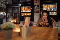 Asian woman smiling and browsing smartphone while sitting at restaurant table near suitcase in evening on city street — Stock Photo