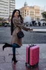 Asian woman with suitcase standing on pavement near road and looking away while visiting city on sunny day — Stock Photo