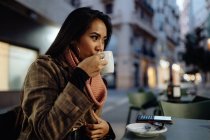 Mujer asiática en ropa de moda sorbiendo bebida caliente fresca y mirando hacia otro lado mientras está sentado en la mesa y descansando en el restaurante de la calle por la noche - foto de stock