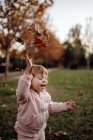 Criança lúdica ativa em roupa quente rosa com olhos fechados do prazer que vomita folhas de outono no prado no parque — Fotografia de Stock