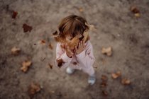 Активна грайлива дитина в рожевому теплому одязі з закритими очима від задоволення підкидає осіннє листя на лузі в парку — стокове фото