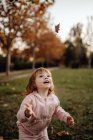Aktives spielerisches Kind in pinkfarbener warmer Kleidung aus Vergnügen, das Herbstlaub auf der Wiese im Park wirft — Stockfoto