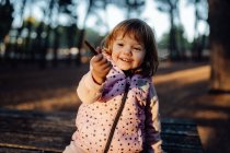 Чарівна активна дитина в теплій рожевій куртці тримає палицю і показує на камеру в сонячному світлі в парку — стокове фото