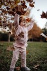 Aktives spielerisches Kind in rosa warmer Kleidung mit geschlossenen Augen vor Vergnügen, das Herbstlaub auf der Wiese im Park wirft — Stockfoto