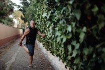 Пожилой мужчина растягивает ногу, держа ботаническую стену на улице. — стоковое фото