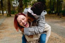 Elegante animado casal multiétnico de jovens namoradas encantadoras se divertindo juntos piggybacking no parque em dia ensolarado — Fotografia de Stock