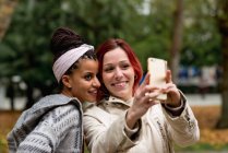 Preciosa pareja multiétnica de mujeres sonrientes que se quedan cerca y se toman selfie en el teléfono móvil en el parque de otoño - foto de stock