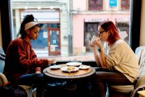 Side view of stylish enthusiastic multiethnic girl friends talking and drinking sitting at table nearby window in cafe — Stock Photo