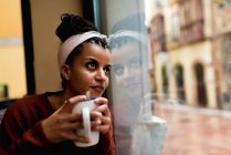 Thoughtful charming African American woman leaning on glass window drinking coffee and dreamily looking away — Stock Photo