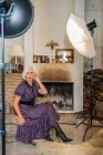 Pensiva atriz de cabelos grisalhos em elegante sentado ao lado de caixa macia e olhando para a câmera durante a pausa no trabalho contra interior borrado de estúdio contemporâneo acolhedor — Fotografia de Stock