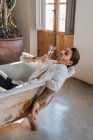 Ernsthafte kühle Barfußrebellin in eleganter Kleidung entspannt sich allein in der Badewanne und feiert den eigenen Erfolg vor rustikalem Interieur im Landhaus — Stockfoto