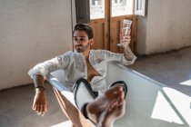 Serious cool barefoot rebel in elegant clothes relaxing alone in bathtub and celebrating own success against rustic interior in country house — Stock Photo