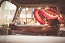 Side view of cheerful young African American woman in colorful wide brimmed striped hat sitting behind wheel of vintage van — Stock Photo