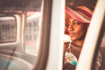 Joven mujer afroamericana positiva en colorido sombrero a rayas sentado detrás del volante de un coche viejo y mirando hacia otro lado - foto de stock