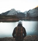 Back view of man in hat and warm jacket with backpack enjoying dark water reflecting sky and rocky snowy mountains around shore — Stock Photo