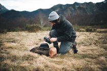 Вид сбоку на человека в шляпе и теплую куртку, снимающего аксессуары с камеры на лугу с сухой травой — стоковое фото