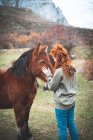 Vista laterale di donna sorridente con lunghi capelli rossi in felpa con cappuccio accarezzando cavallo marrone con criniera nera in pascolo di montagna — Foto stock