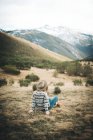 Back view of unrecognizable pensive charming woman with red hair in warm sweater sitting on lawn in mountain in gray day — Stock Photo