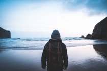 Back view of anonymous traveler in jacket with backpack contemplating views of seaside surrounded with mountain — Stock Photo