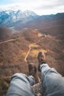 Viajante de culturas em botas marrons sentado com as pernas penduradas na borda da rocha e desfrutando de uma vista deslumbrante da floresta e da estrada — Fotografia de Stock