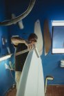 Qualified worker in uniform handling white surfboard in workshop with blue walls — Stock Photo
